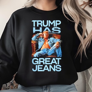 May include: Black sweatshirt with the text "TRUMP HAS GREAT JEANS" in light blue. The graphic depicts a person in a denim jacket and jeans. The person is seated in a brown chair.