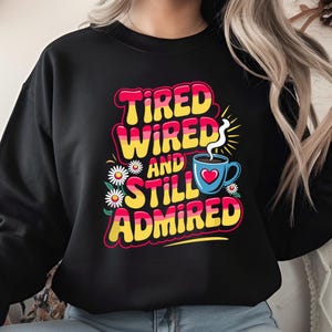Può includere: Felpa nera con un design grafico colorato che dice "Tired Wired and Still Admired" con una tazza di caffè fumante e fiori.