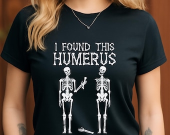 Peut inclure: T-shirt noir avec l'inscription blanche "I FOUND THIS HUMERUS". Le motif présente deux squelettes et un os du bras détaché. Un design humoristique pour Halloween ou les amateurs de science.