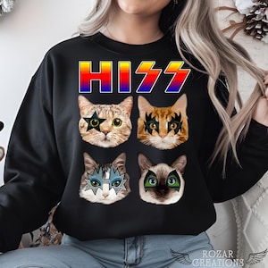 Pode incluir: Camisola preta com a palavra "HISS" em cores do arco-íris e quatro rostos de gato com maquiagem de estrela do rock. Os gatos usam óculos de sol e têm um tema de rock and roll.