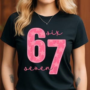 May include: Black t-shirt with a pink graphic featuring the numbers 6 and 7, filled with white stars. The word "six" is above the number 6, and "seven" is below the number 7, in a cursive font. The shirt has a crew neck.