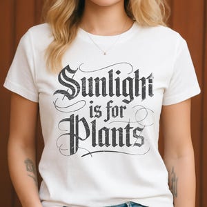 Puede incluir: Camiseta blanca con la frase "Sunlight is for Plants" en fuente negra desgastada. La camiseta tiene mangas cortas y cuello redondo. El texto está en una fuente decorativa de estilo vintage.