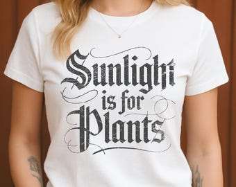 Puede incluir: Camiseta blanca con la frase "Sunlight is for Plants" en fuente negra desgastada. La camiseta tiene mangas cortas y cuello redondo. El texto está en una fuente decorativa de estilo vintage.