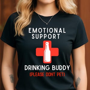 May include: Black t-shirt with the words "EMOTIONAL SUPPORT" above a red cross with a white bottle graphic. Below the cross, the text reads "DRINKING BUDDY" and "(PLEASE DONT PET)".