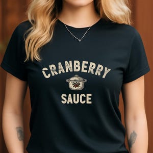Puede incluir: Camiseta negra con la frase "CRANBERRY SAUCE" en una fuente desgastada de color blanquecino. Un gráfico de una olla con un diseño floral se encuentra entre las palabras. La camiseta es de cuello redondo.