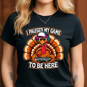 May include: Black t-shirt featuring a cartoon turkey wearing headphones and a hat, holding a game controller. The text reads "I PAUSED MY GAME TO BE HERE" in white and orange. The turkey has a red wattle and a brown body.
