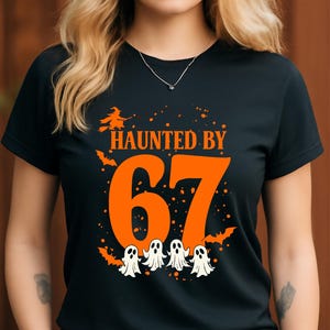 Puede incluir: Camiseta negra con el texto naranja "HAUNTED BY 67". El diseño incluye una bruja, murciélagos y cuatro fantasmas blancos. Una camiseta gráfica con temática de Halloween.