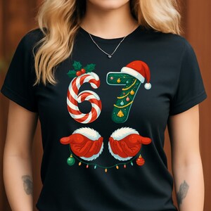Op de afbeelding: Zwart T-shirt met een feestelijk kerstontwerp. Het ontwerp bevat de cijfers 6 en 7, een zuurstok, een kerstboom en handen die ornamenten vasthouden. Het shirt heeft een ronde hals.