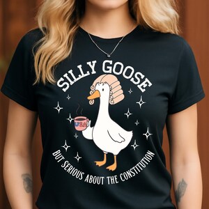 Op de afbeelding: Zwart T-shirt met een afbeelding van een witte gans met een rechters pruik en een koffiekopje met "USA". De tekst luidt "SILLY GOOSE BUT SERIOUS ABOUT THE CONSTITUTION" met steraccenten.
