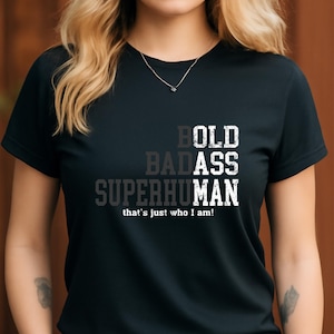 Può includere: T-shirt nera con la scritta "BOLD BADASS SUPERHUMAN" in un design a strati. La frase "that's just who I am!" è stampata sotto. La maglietta ha un girocollo e maniche corte.