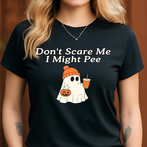 Può includere: T-shirt nera con la scritta "Don't Scare Me I Might Pee". La maglietta presenta un fantasma dei cartoni animati con un berretto arancione e che tiene in mano una zucca e una bevanda. Una divertente maglietta grafica a tema Halloween.