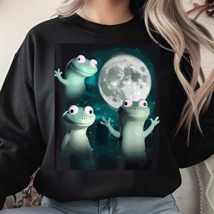 Può includere: Felpa nera con un'illustrazione stravagante di tre rane verdi con occhi rosa, ambientata su uno sfondo blu scuro stellato e una grande luna piena. Le rane sono raffigurate con le braccia alzate.