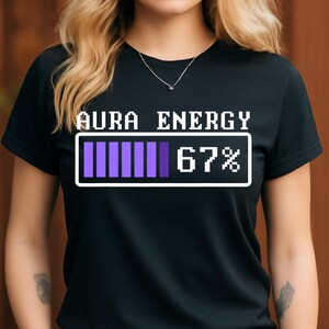 May include: Black t-shirt with the text "AURA ENERGY" above a pixelated purple energy bar showing 67% charge. The shirt has a crew neck and short sleeves.