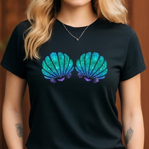 May include: Black t-shirt featuring a glittery seashell design in shades of teal, blue, and purple. The graphic is centered on the front of the shirt. The shirt has short sleeves and a crew neck.