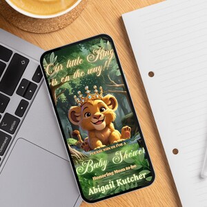 May include: A smartphone displays a baby shower invitation. The invitation features a cartoon lion cub wearing a crown, with text that reads "Our little King is on the way!!" and "Baby Shower". The invitation is for Abigail Kutcher.