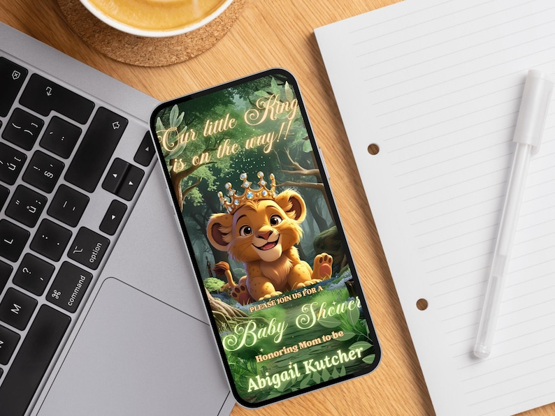 May include: A smartphone displays a baby shower invitation. The invitation features a cartoon lion cub wearing a crown, with text that reads "Our little King is on the way!!" and "Baby Shower". The invitation is for Abigail Kutcher.