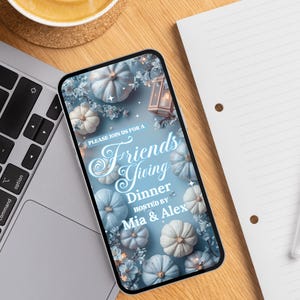 May include: A smartphone displays a Friendsgiving dinner invitation. The invitation features a light blue background with white and blue pumpkins, floral accents, and the text "PLEASE JOIN US FOR A Friends Giving Dinner HOSTED BY Mia & Alex."