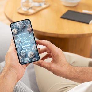 May include: A smartphone displays a Friendsgiving dinner invitation. The invitation features a design with light blue pumpkins and floral accents. The text reads "Friendsgiving Dinner" hosted by Mia & Alex.