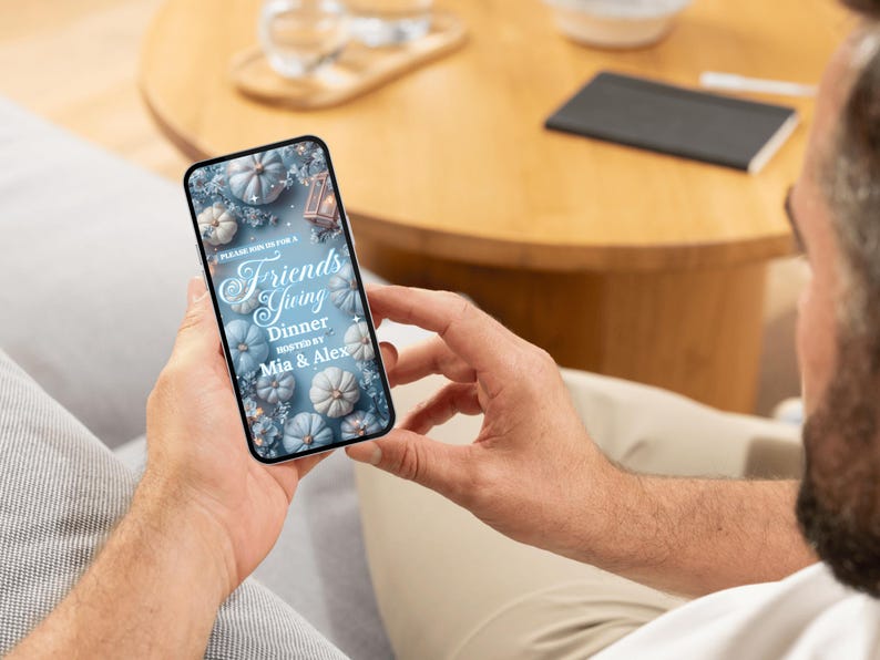 May include: A smartphone displays a Friendsgiving dinner invitation. The invitation features a design with light blue pumpkins and floral accents. The text reads "Friendsgiving Dinner" hosted by Mia & Alex.