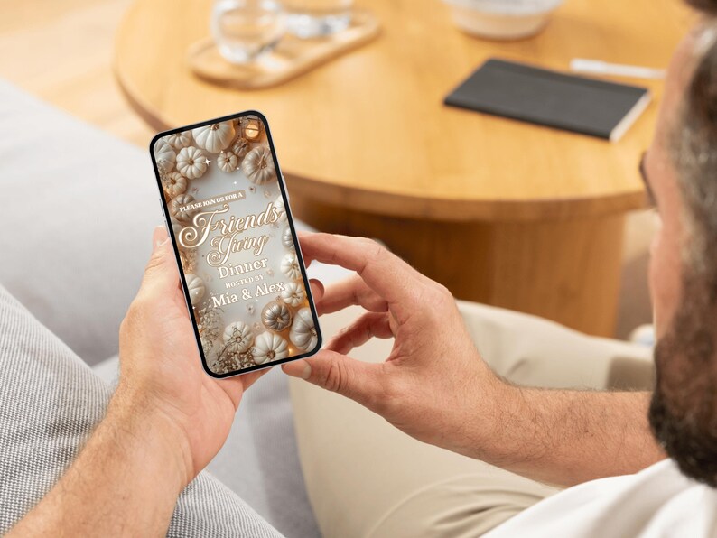 May include: A smartphone displays a Friendsgiving dinner invitation. The design features white and gold pumpkins, leaves, and text. The invitation includes the words "Friendsgiving Dinner" and the names "Mia & Alex."