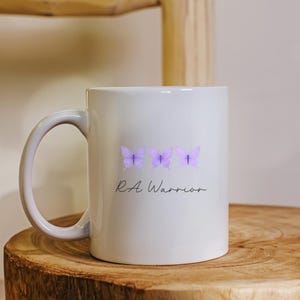 May include: A white ceramic mug featuring a horizontal row of three light purple butterflies printed on the front. Below the butterflies, the text RA Warrior is written in a black, cursive script font. The mug has a curved handle on the left side.
