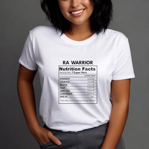 May include: A white short-sleeved t-shirt featuring a black graphic print of a Nutrition Facts label. The text reads RA WARRIOR, Serving Size: 1 Super Hero, and lists attributes like STRENGTH, COURAGE, BELIEVE, FIGHT, AMBITION, and CONFIDENCE at 1000%.