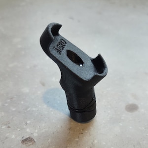 May include: A black 3D-printed object with the text "AS3D" on the top. The object has a curved top with a hole in the centre and a cylindrical base. The base is wrapped with a thin black wire.