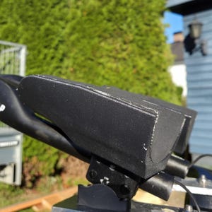 May include: Black automotive part with the text "51 SPEEDSHOP" on a curved arm. The part has a rectangular shape with rounded edges and is mounted on a metal base. The background includes a green bush and a blue building.