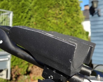 May include: Black automotive part with the text "51 SPEEDSHOP" on a curved arm. The part has a rectangular shape with rounded edges and is mounted on a metal base. The background includes a green bush and a blue building.