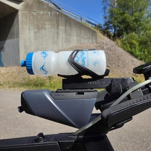Puede incluir: Una botella de agua para bicicleta con tapa azul y un portabotellas negro y gris montada en una bicicleta. La botella tiene un diseño de delfín azul y la palabra "Salish". La bicicleta es negra y gris.