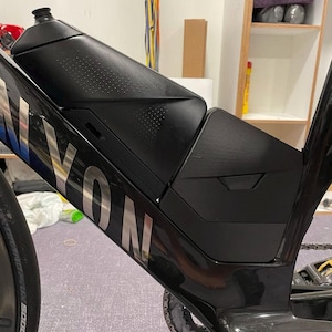 Puede incluir: Bicicleta de carretera Canyon negra con una botella de agua y un compartimento de almacenamiento. El cuadro de la bicicleta es negro con letras plateadas. La botella de agua es negra y tiene una superficie texturizada.