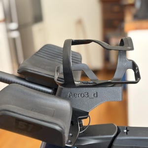 May include: Black bicycle water bottle cage with the text "Aero3_d" attached to a bicycle. The cage is designed to hold a water bottle and is mounted on the handlebars. The image also shows a black armrest.
