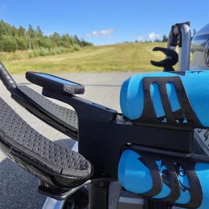 May include: Close-up of a black and blue triathlon bike. The bike has a black frame, blue water bottles, and a phone mount. The handlebars have textured grips. The background shows a blue sky and green grass.
