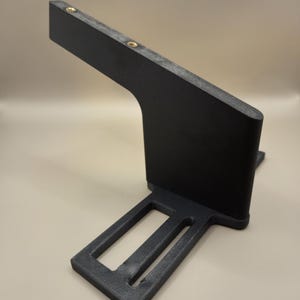 Op de afbeelding: Een zwarte, schuine beugel met twee messingkleurige schroeven. De beugel heeft een rechthoekige basis met twee uitgesneden secties en een gebogen steunarm. Het object is gemaakt van een mat materiaal.