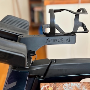 May include: Black bicycle accessory with the text "Aero3_d" on the side. The accessory includes a water bottle cage and armrests. The item is mounted on a bicycle and appears to be made of plastic.