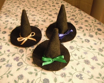 May include: Three black felt witch hats with pointed crowns and wide brims. Each hat features a small satin ribbon tied in a bow around the base of the crown. One hat has a gold ribbon, one has a purple ribbon, and one has a green ribbon.
