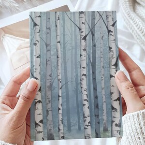 May include: A rectangular notebook featuring a printed illustration of a birch forest. The cover displays tall, slender white and black birch tree trunks set against a soft, muted blue and grey misty background, creating a layered woodland scene.