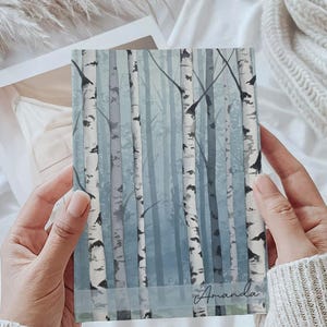 May include: A rectangular notebook featuring a printed forest scene with white birch trees against a muted blue background. A semi-transparent horizontal band across the lower portion displays the name Amanda in a black cursive script font.