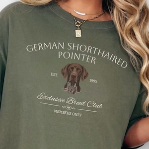 Pode incluir: Camiseta verde-oliva com a inscrição "GERMAN SHORTHAIRED POINTER" e uma ilustração de um cão. Abaixo da imagem do cão, o texto diz "Exclusive Breed Club" e "MEMBERS ONLY". A camiseta também tem o ano de "1995".