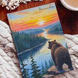 May include: A journal with a landscape illustration. The cover depicts a bear overlooking a lake and forest at sunset. The name "Amanda" is written in cursive at the top. The colour palette includes shades of orange, blue, and brown.