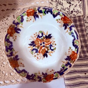 Puede incluir: Un plato de cerámica blanca con borde festoneado, adornado con un patrón floral en tonos naranja, azul y verde. El plato presenta un ramo floral central y motivos florales repetidos alrededor del borde, creando una estética vintage.