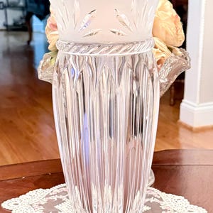 May include: A clear glass vase with vertical ribbed detailing. The top section features a frosted design with leaf patterns. A decorative rope-like band encircles the vase's midsection. The vase is displayed on a wooden table with a lace doily.