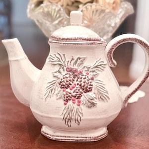 May include: Cream-colored ceramic teapot with a lid and handle. The teapot features a raised design of pinecones and leaves, with accents of brown and pink. The spout and handle are also brown. The teapot is sitting on a wooden surface.