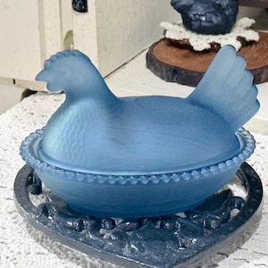 May include: A blue glass hen-shaped covered dish. The lid is shaped like a hen with detailed feathering. The dish sits on a dark metal base with a floral design. The dish is a light blue color.