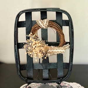 May include: Black rectangular basket with a woven design, holding a small wreath. The wreath is made of brown twigs, dried flowers, and cream-colored lace. The basket is on a black metal stand.
