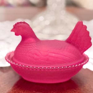 May include: A vibrant pink hen-shaped trinket box. The lid features a detailed hen with a comb and tail feathers. The base is a bowl shape, adorned with a white beaded trim. A decorative item for the home.