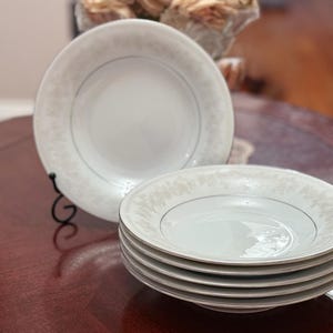 May include: A stack of white porcelain soup bowls with a delicate floral pattern and a silver rim. One bowl is displayed on a black metal stand. The bowls are on a dark wood table.
