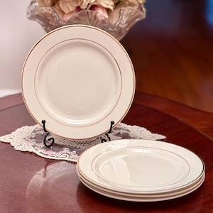 Puede incluir: Platos de cerámica blanquecina con borde dorado. Un plato se exhibe sobre un soporte de metal negro, mientras que otros tres están apilados sobre una superficie de madera oscura. Un tapete de encaje y un jarrón de cristal con flores están en el fondo.