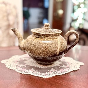 Può includere: Una teiera in ceramica marrone e beige con coperchio, beccuccio e manico. La teiera è appoggiata su un centrino di pizzo bianco con accenti floreali rosa. La teiera ha una superficie strutturata e una base marrone scuro.