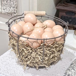 Op de afbeelding: Een draadmand gevuld met lichtroze eieren en versnipperd papier. De mand heeft een houten handvat en een metalen frame. De eieren zijn glad en gelijkmatig gekleurd, wat wijst op een decoratief doel.